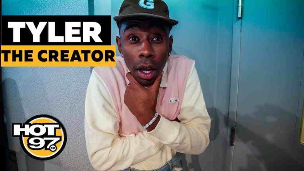 American Rapper Tyler The Creator Hand holding chin with arms folded wearing a cap with a G on it with a pink and white shirt